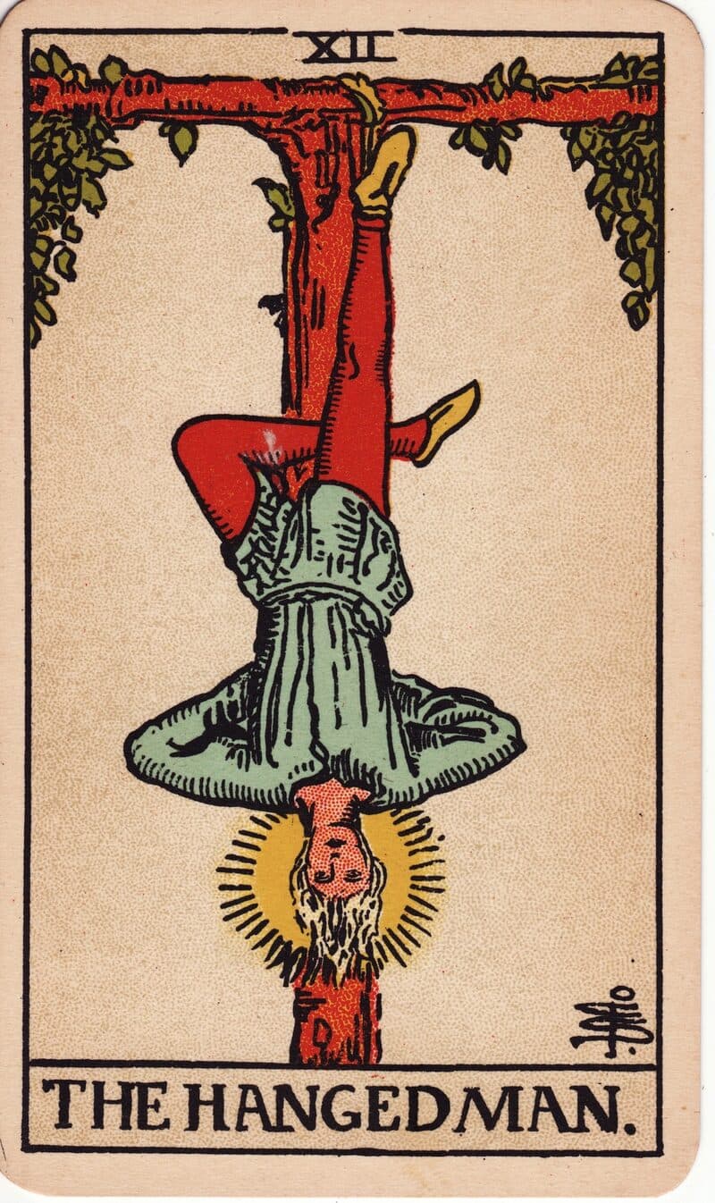 Carta de El Colgado — figura suspendida cabeza abajo de una cruz tau con halo de luz, baraja Rider-Waite-Smith