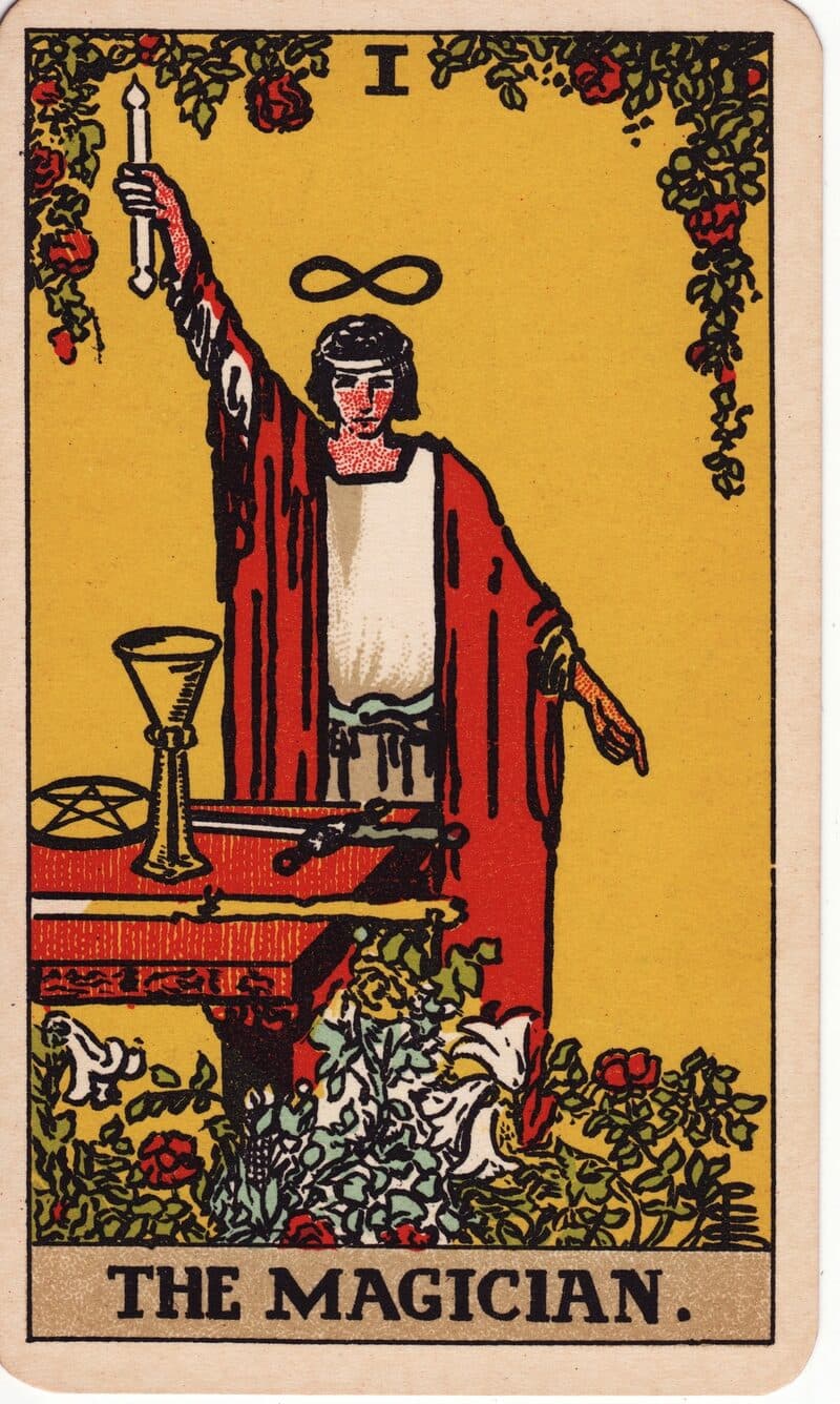 Carta de El Mago — figura con túnica ante altar con los cuatro emblemas y una mano alzada, baraja Rider-Waite-Smith